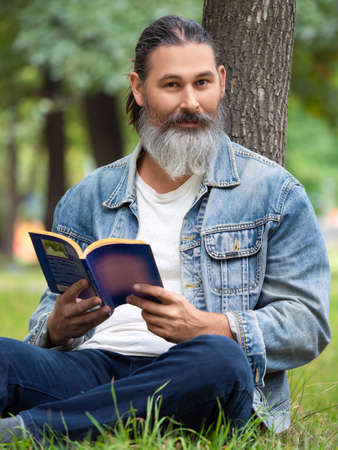 Vertical photo of a middle aged man with a book in the park. He sits on the grass in the park and looking at camera. Digital detox concept.の写真素材