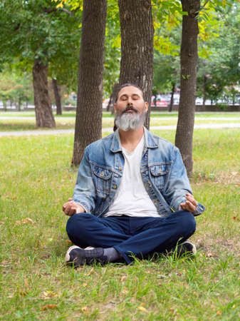Vertical photo of a bearded middle-aged man taking a break from the hustle and bustle of the city. He is meditating in the park under a tree. Digital detox concept.の写真素材