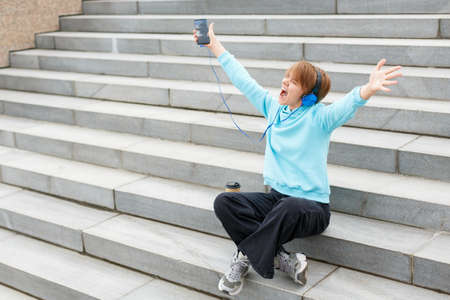 Young hipster girl sits on the stairs and listens to music with her arms outstretched. She listens...
