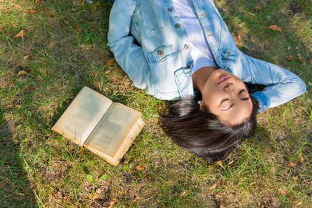 Digital detox concept. Young Asian woman relaxing with a book - she lying on the grass in city park.の写真素材