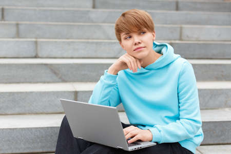 Photo of a young woman with short hair is sitting on stairs witl laptop. Online education concept.の写真素材