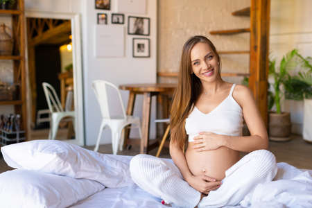 A young pregnant woman is sitting on the bed - she waking up, looking at camera and smiling.の写真素材