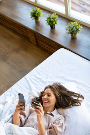 Woman lying in bed makes an order through a mobile application. She dials the card number for online purchases. Top view.の写真素材