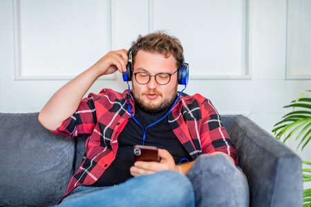 Photo of a young man works at home sitting on sofa in headphones and with smartphone. He is listening to the music via online app for smartphones.の写真素材