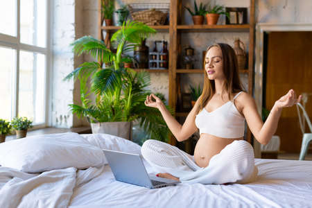 A pregnant woman is meditating in front of a laptop monitor while sitting on the bed.の写真素材