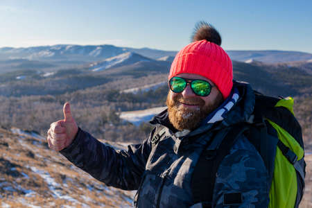 Bearded smiling male tourist in sunglasses keeping thumb up on the top of the mountain - he posing for photo.の写真素材
