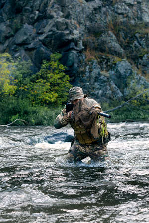 Vertical photo of a mercenary military soldier during a sabotage mission behind enemy lines - he stands knee-deep in the river and aims at the enemy.の写真素材