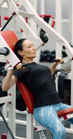 Slow motion 4k footage. In the gym, a middle-aged woman uses the hammer machine for bench presses,...