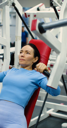 Gym workout. Bench press in hammer machine. Caucasian woman trains her chest muscles with focused...