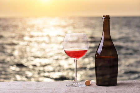 Bottle and glass of red wine on the linen table against the sea or ocean on sunsetの写真素材