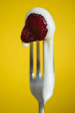 Macro shot of ripe strawberries on a fork with sour cream. Closeup red berry with yellow backgroundの写真素材