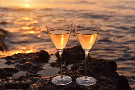 Two glasses with white wine on sthe stone near the sea. Golden sunset