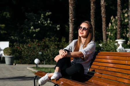 young woman sitting on bench in park, smiling and looking into the camera at sunsetの写真素材