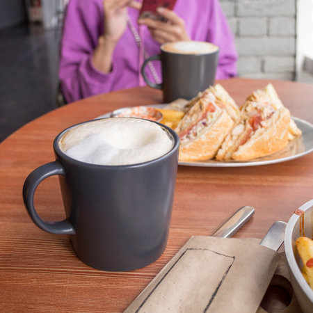 black coffee mug with white foam on the table in a cafe against the background of a woman with a smartphone. copyspace.の写真素材