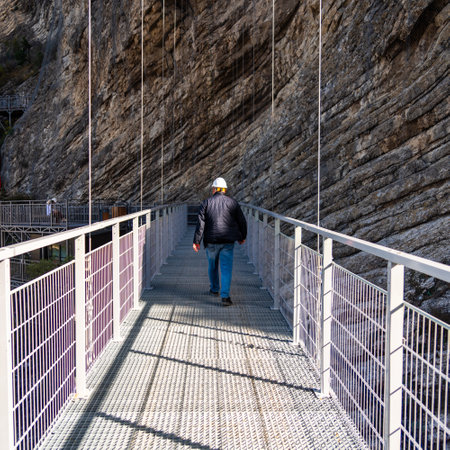 a man in a protective construction helmet walks along a suspended metal bridge in the mountains...