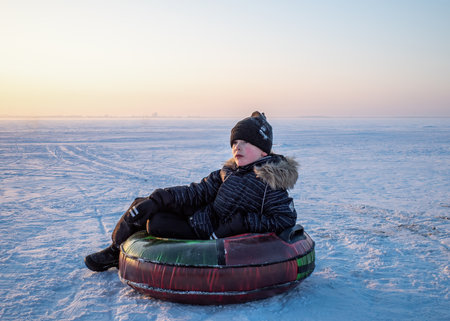young boy dressed in winter clothing sits on an inflatable snow tube on a vast, snow-covered plain under a pale sky with a distant city skyline. natural lightの写真素材