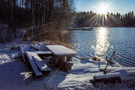 snow-covered picnic table and benches by a lake, with the sun reflecting brilliantly on the waterの写真素材