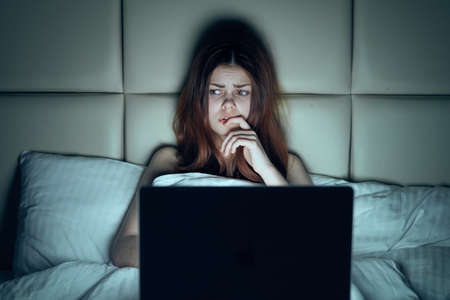 The thoughtful woman sits in bed with a laptop.の写真素材