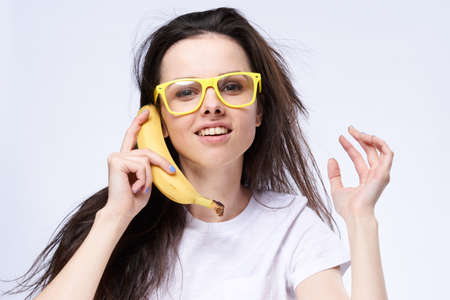 A woman in glasses holds a banana near the ear on a light background.の写真素材