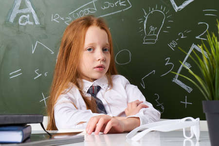 Schoolgirl sitting at the table, on the table glasses, schoolgirl on the background of the school board.の写真素材