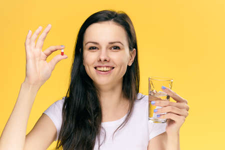 Woman with a pill and a glass of water on a yellow background.