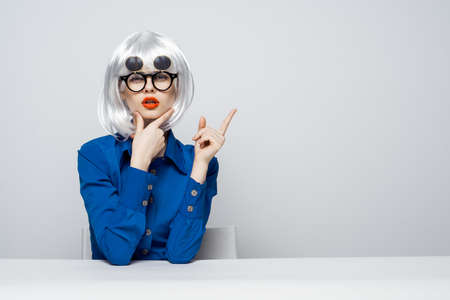 Woman in a wig, woman in funny glasses, a young woman sits at a table on a light background.