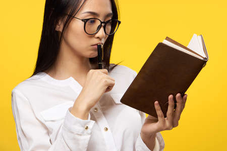 Female student with glasses, a young woman looks at a notepad, a woman is holding a pen on a yellow background.の写真素材