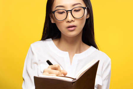 Asian, girl on a yellow background writes in a notebook, woman in glasses, student, teacher.の写真素材