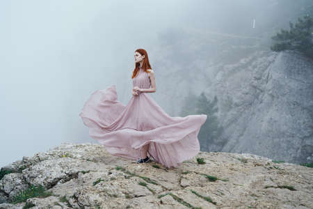 Beautiful young woman in a long dress is standing on a cliff of a mountain.の写真素材
