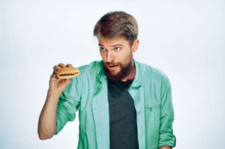 Young guy with a beard on a light background holds a hamburger.