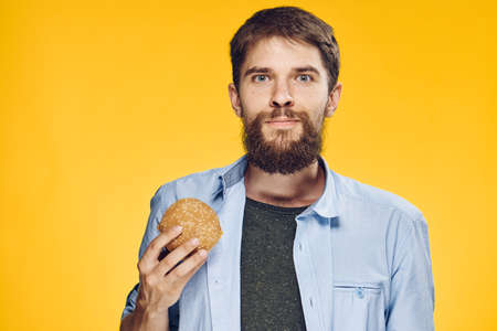 Young guy with a beard on a yellow background holds a hamburger.