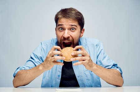 Young guy with a beard on a light background holds a hamburger.
