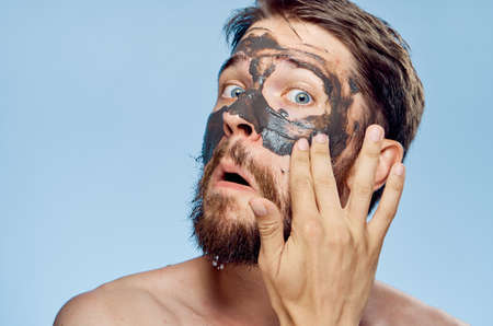 A young guy with a beard on a blue background applies clay cosmetic face mask.の写真素材