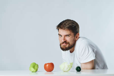 Man with a beard on a white isolated background sits at a table and looks at vegetables, diet, vegetarianism, vegetarian.の写真素材