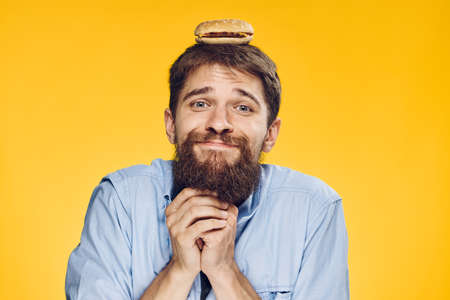 Man with a beard on a yellow background holds a hamburger on his head.の写真素材