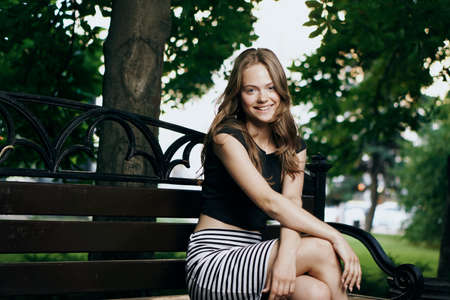 Young beautiful woman sits on a summer bench in a park in the city.の写真素材