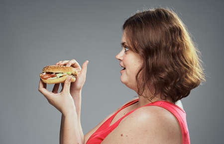 Fat woman on a gray background holds a hamburger, fast food.の写真素材
