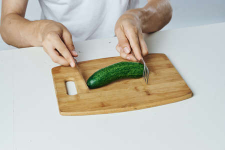 Man at the table on a white isolated background cuts a cucumber on a cutting board, vegetarianism, diet.の写真素材