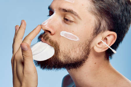 A man with a beard on a blue background holds wadded disks in his mouth, cosmetic cream, ear wand.の写真素材