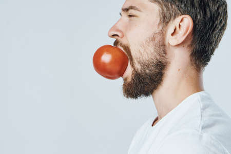 Man with a beard on a white isolated background holds a tomato in his mouth, vegetables, vegetarian, diet.の写真素材
