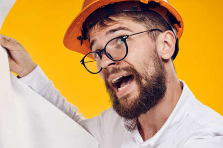 Engineer with a beard in a helmet on a yellow background holds blueprints, portrait, construction.の写真素材