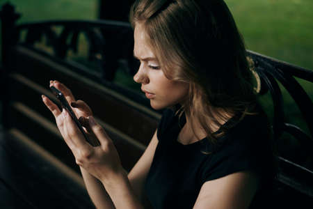 Young beautiful woman is sitting in the phone on a park bench in summer.の写真素材
