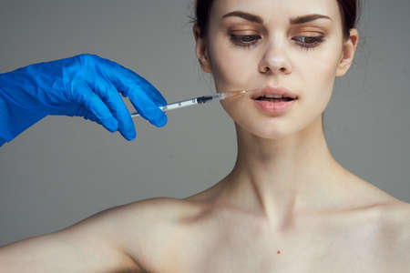 Young beautiful woman on a gray background in rubber medical gloves holds a syringe, medicine,...