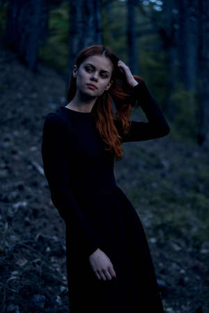 Young beautiful woman in a black long dress is standing in a dark forest.の写真素材