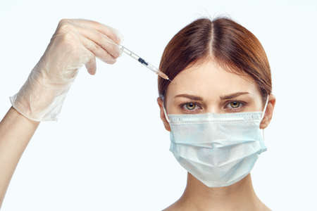Young beautiful woman on a light background in a medical mask holds a syringe, medicine, plastic surgery.の写真素材