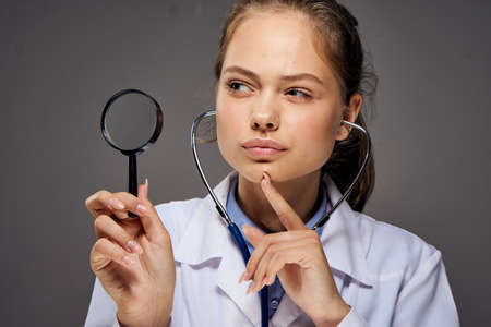 Young beautiful woman on a gray background in a medical dressing gown holds a magnifier, doctor, medicine.の写真素材