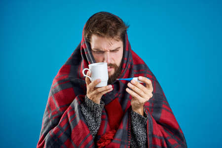 A man with a beard on a blue background holds a mug and a thermometer in a plaid, sickness, sick.の写真素材