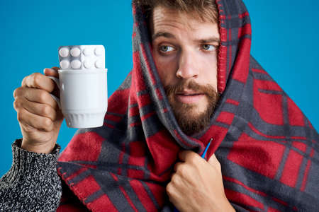 A man with a beard holding pills and a mug in a plaid on a blue background, emotions, a portrait, sick, a disease, a cold.の写真素材