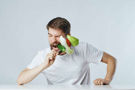 Man with a beard on a light background sits at a table and keeps on a fork vegetables, vegetarian, diet, emotions.の写真素材