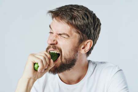 Man with a beard on a light background bites off a cucumber, portrait.の写真素材